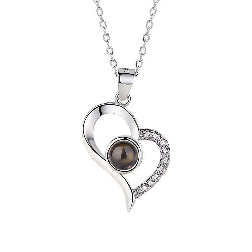 A silver necklace with a heart-shaped pendant featuring a central dark stone and surrounding small clear stones along the perimeter.