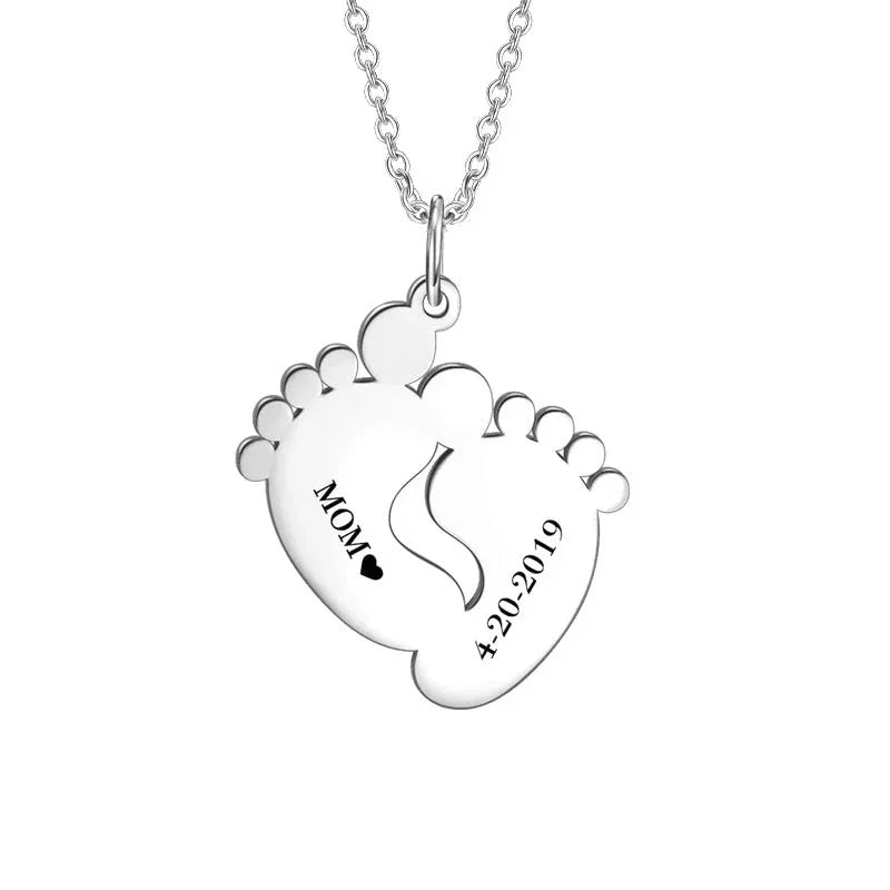A silver necklace featuring a pendant shaped like two overlapping baby feet. One foot is engraved with the word “MOM” followed by a small heart symbol, and the other displays the date “4.20.2019.” The pendant hangs from a delicate silver chain against a plain white background.