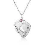 Silver necklace with a pendant shaped like two baby feet forming a heart, engraved with the name “Lucy” and date “12.05.20,” featuring a small pink birthstone.