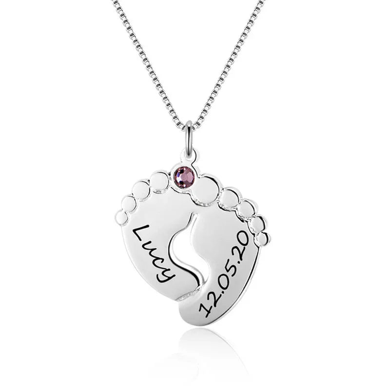 Silver necklace with a pendant shaped like two baby feet forming a heart, engraved with the name “Lucy” and date “12.05.20,” featuring a small pink birthstone.