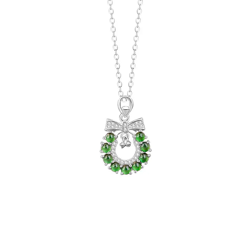 A delicate silver necklace featuring a circular Christmas wreath pendant. The wreath is adorned with jade-like gemstones and accented with a small bell and a bow at the top, creating a festive and elegant holiday design.