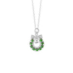 A delicate silver necklace featuring a circular Christmas wreath pendant. The wreath is adorned with jade-like gemstones and accented with a small bell and a bow at the top, creating a festive and elegant holiday design.