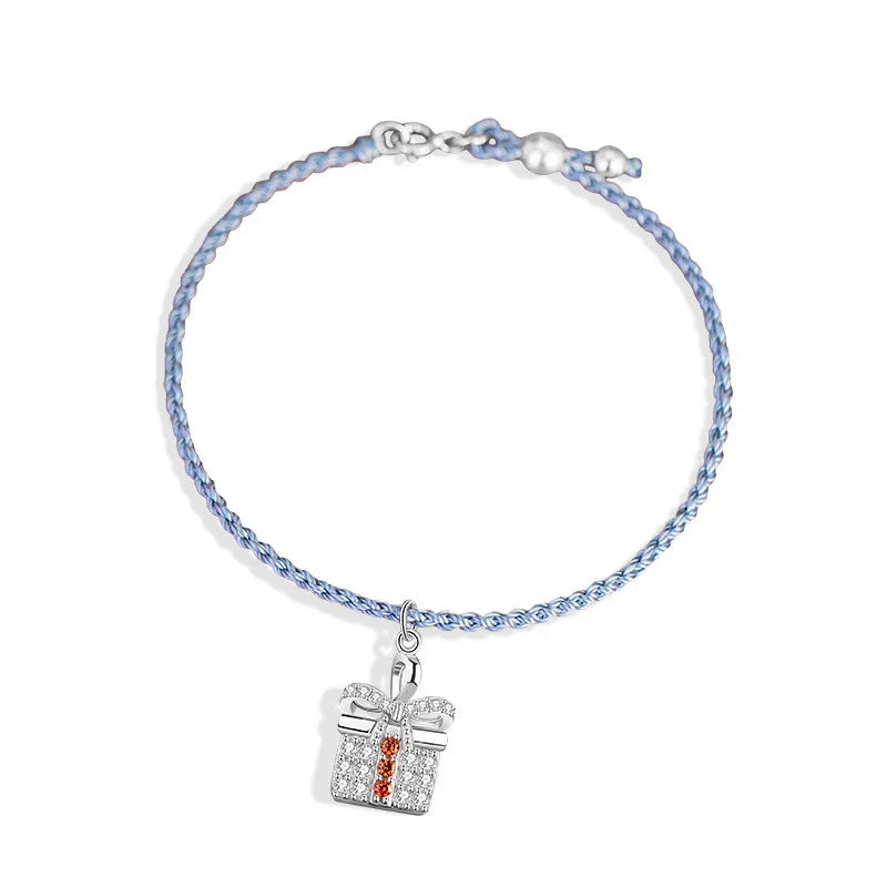 A delicate Christmas-themed bracelet featuring a braided light blue cord, accented with small silver beads, and a festive gift box charm adorned with sparkling clear stones and a vertical row of red gems.