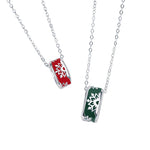 A pair of silver couple necklaces featuring cylindrical pendants with snowflake cutout designs. One pendant is decorated with a festive red background, while the other has a green background, both hanging from delicate silver chains and displayed on a white background.