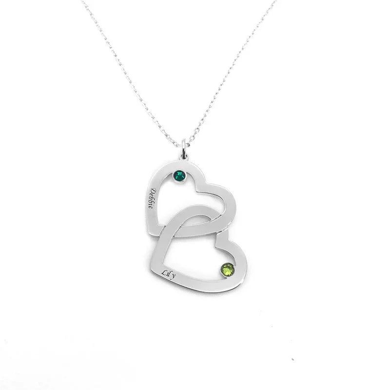 A silver necklace featuring two interlocking heart pendants, each engraved with a name and accented with a small green gemstone, symbolizing love and connection.