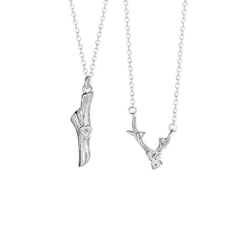 A pair of silver couple necklaces displayed on a white background. One pendant features a straight tree branch with a small heart-shaped gemstone, while the other resembles a forked branch with a round sparkling stone. Both are attached to delicate silver chains, symbolizing connection and love.