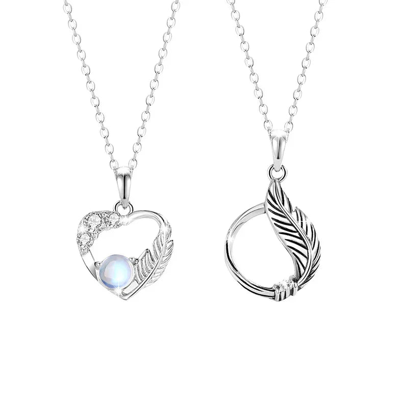 A pair of sterling silver couple necklaces. One features a delicate heart-shaped pendant adorned with small crystals and a luminous gemstone, accented with a feather motif. The other has a circular pendant wrapped with a feather design. Both are displayed on fine silver chains against a white background.