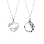 A pair of sterling silver couple necklaces. One features a delicate heart-shaped pendant adorned with small crystals and a luminous gemstone, accented with a feather motif. The other has a circular pendant wrapped with a feather design. Both are displayed on fine silver chains against a white background.