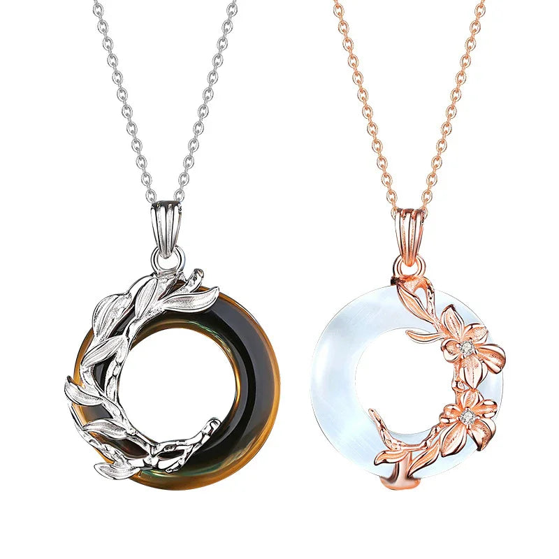 A pair of couple necklaces displayed on a white background. The left necklace features a circular amber-colored pendant wrapped with silver leaves, while the right necklace has a white circular pendant adorned with rose gold flowers and leaves. Both pendants hang from delicate matching chains, symbolizing harmony and love.