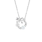 A delicate silver necklace featuring a circular wreath pendant adorned with a small bow, star, snowflake accents, sparkling crystals, and a single white pearl, displayed on a white background.