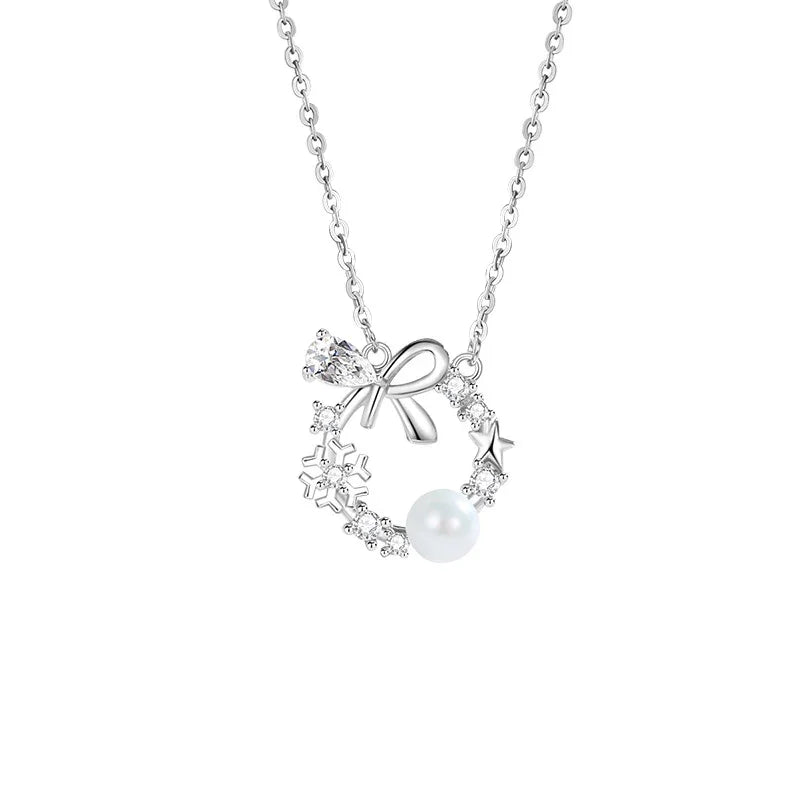 A delicate silver necklace featuring a circular wreath pendant adorned with a small bow, star, snowflake accents, sparkling crystals, and a single white pearl, displayed on a white background.