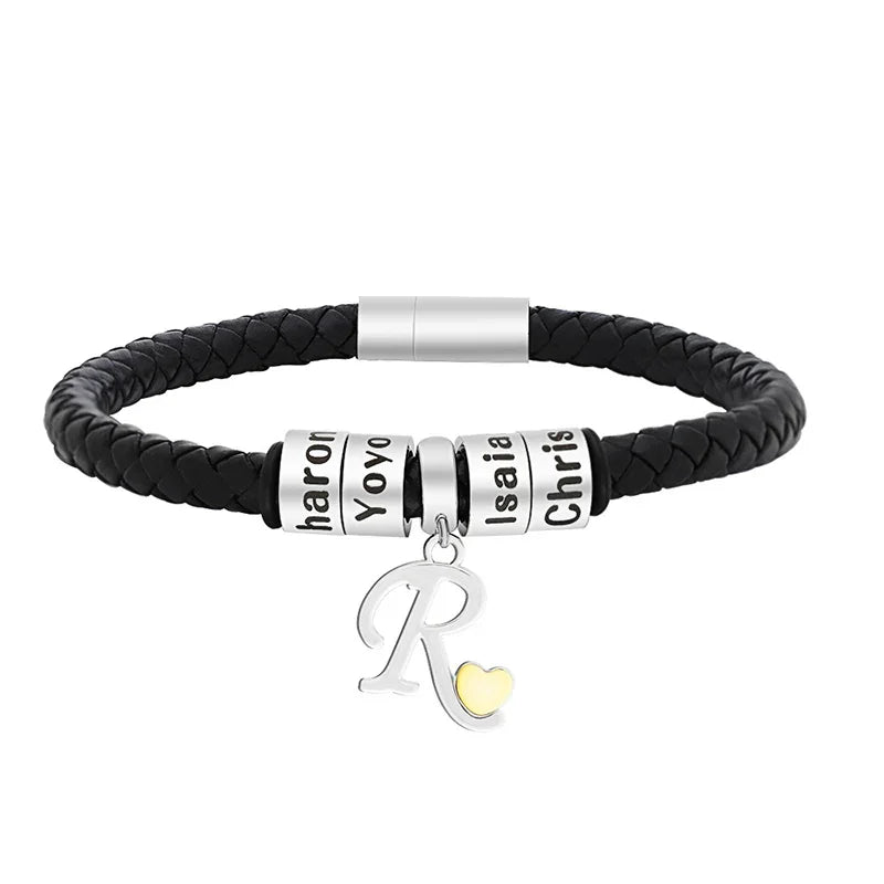 A men's black braided leather bracelet featuring multiple stainless steel name rings engraved with different names and a dangling silver letter “R” charm with a small gold heart accent. The bracelet has a sleek magnetic clasp and a modern, personalized design.