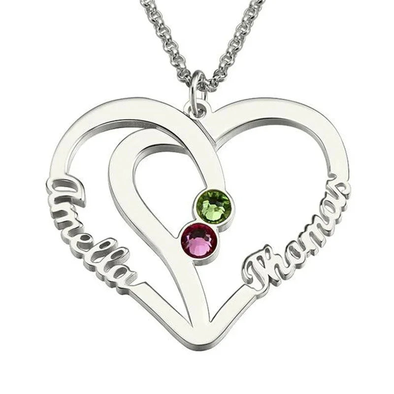 A silver heart-shaped necklace pendant featuring two engraved names, “Amelia” and “Thomas,” with two colorful birthstones — one green and one pink — set in the center, symbolizing love and connection.