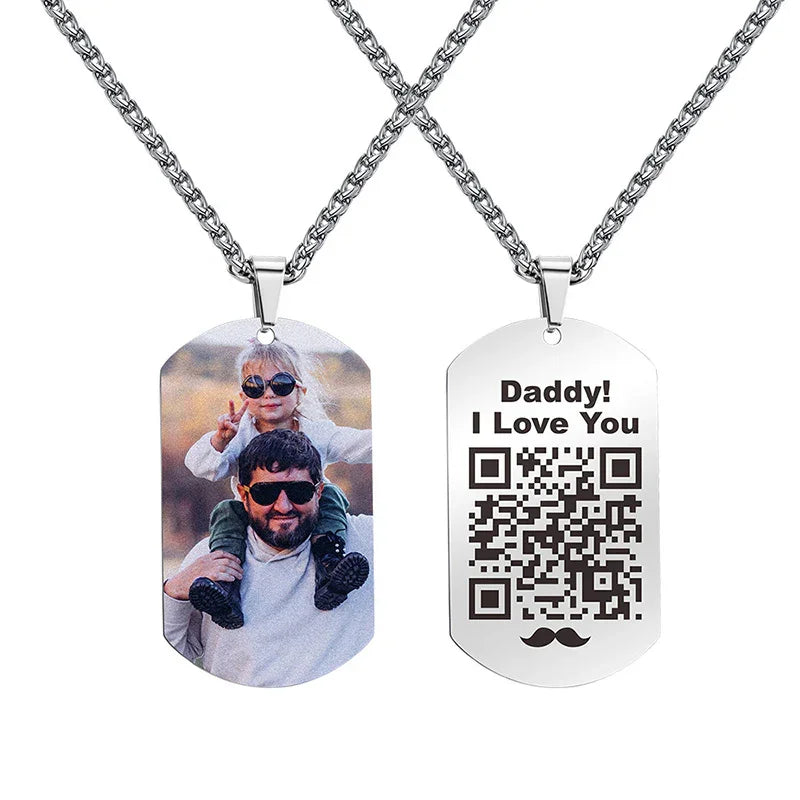 Two stainless steel dog tag necklaces on chains — one engraved with a photo of a father carrying his child on his shoulders, both wearing sunglasses, and the other engraved with the words “Daddy! I Love You,” a QR code, and a small mustache icon.