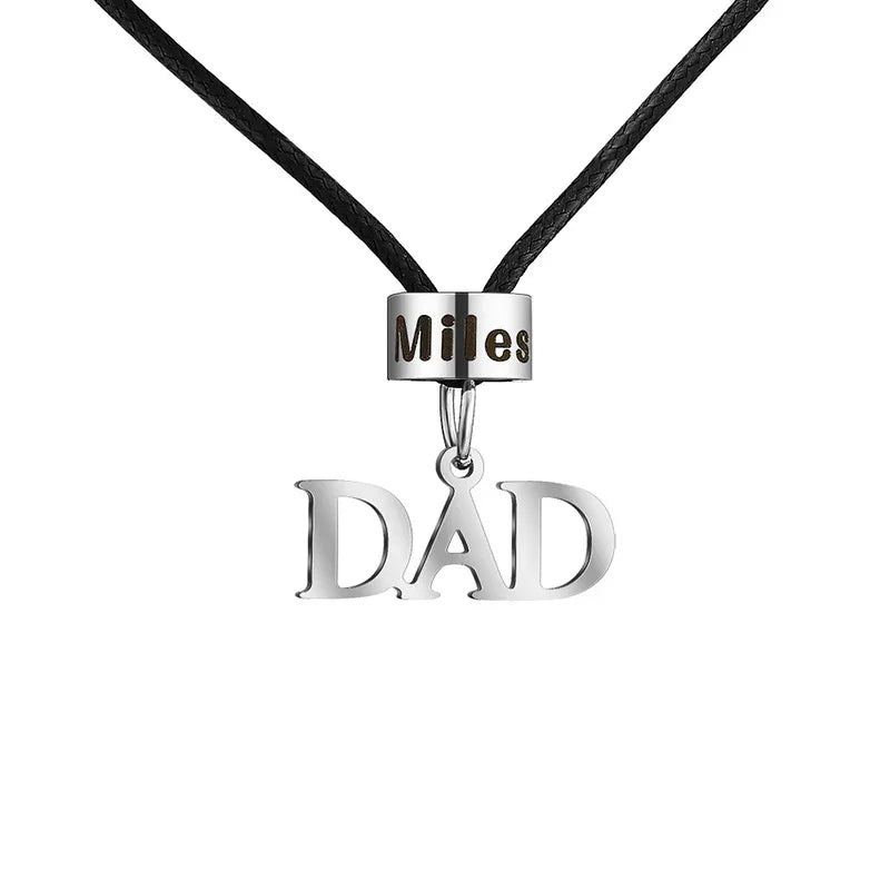 A men’s necklace with a black cord featuring a silver bead engraved with the name “Miles” and a hanging “DAD” pendant.