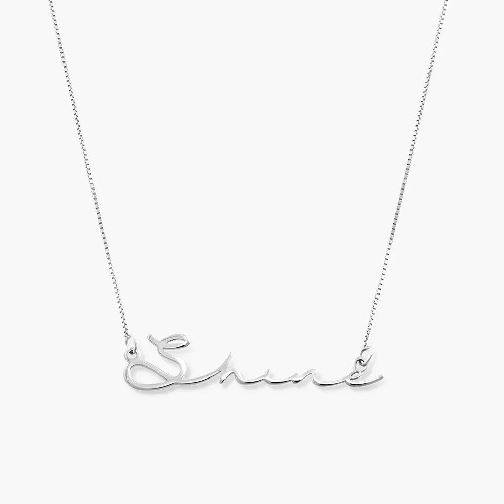 A silver signature-style name necklace featuring the word “Shine” in elegant cursive lettering, attached to a delicate silver chain on both sides. The design is sleek and minimal, highlighting the flowing script of the name.