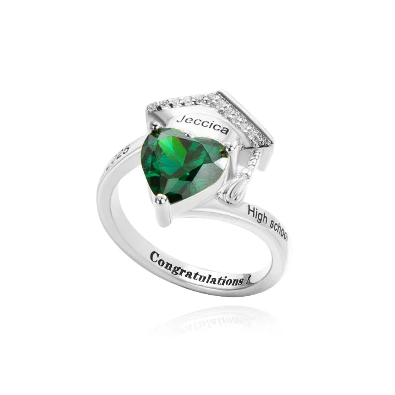 A silver ring featuring a green heart-shaped gemstone set beneath a graduation cap design with engraved details.