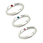 Three slim silver rings with engraved names and small birthstones in red, blue, and pink.