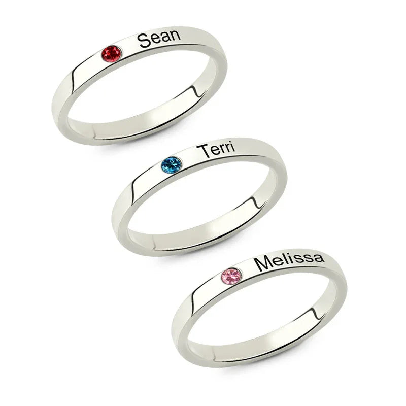 Three slim silver rings with engraved names and small birthstones in red, blue, and pink.