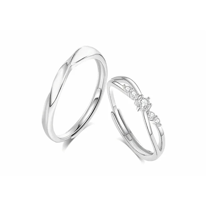 A pair of elegant silver couple rings displayed on a white background. One ring features a smooth, twisted band with a minimalist design, while the other has a delicate crisscross shape adorned with small sparkling gemstones, symbolizing love and unity.