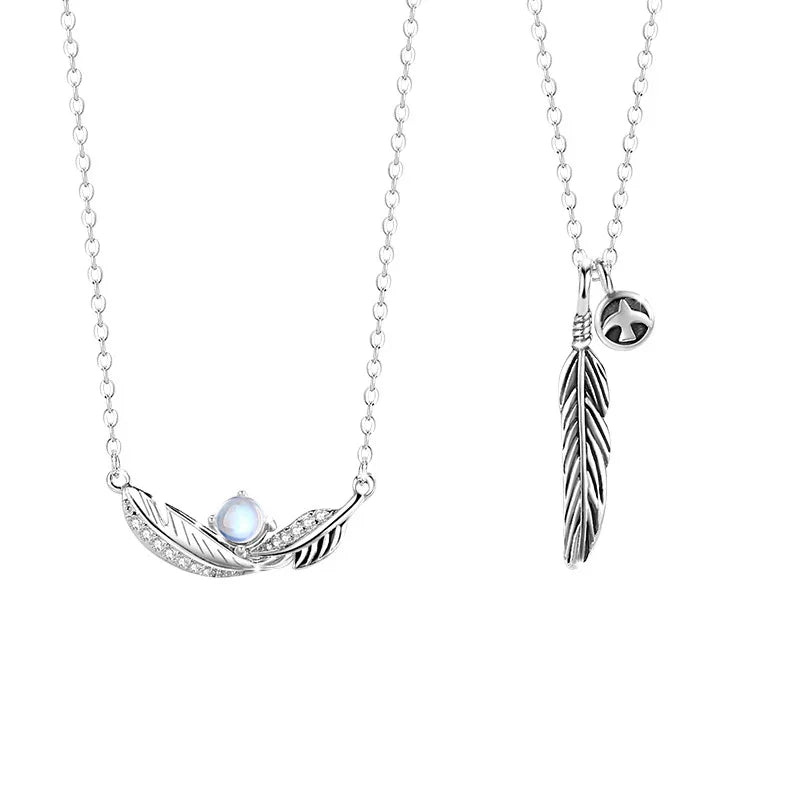 A pair of silver couple necklaces featuring feather pendants — one with a curved feather design adorned with a luminous gemstone and the other with a vertical feather charm and a small star accent, displayed on a white background.